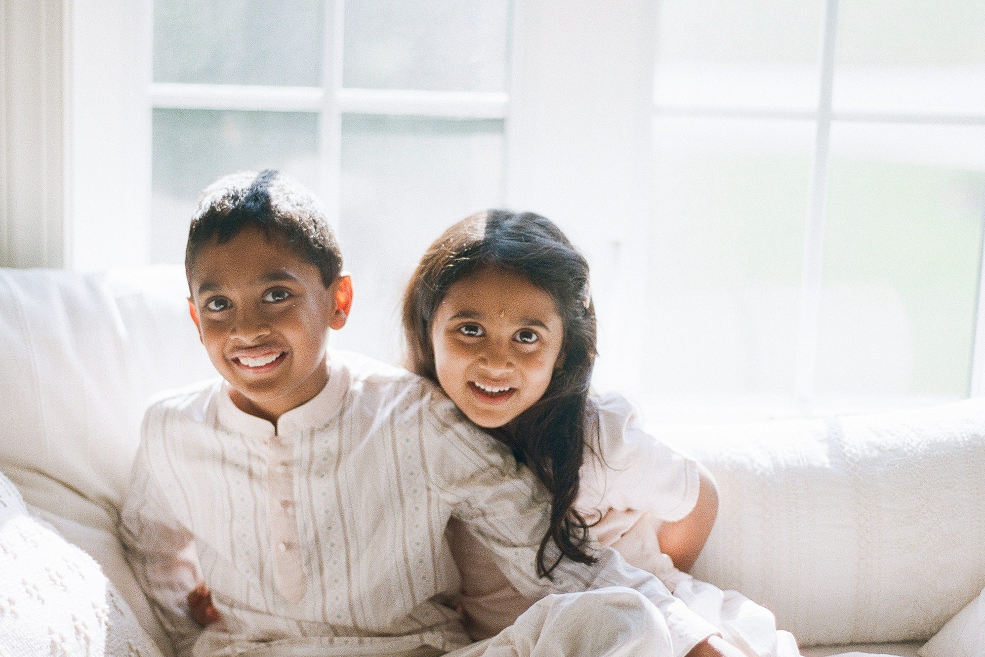 Two children sitting on a white couch in a bright room with large windows.