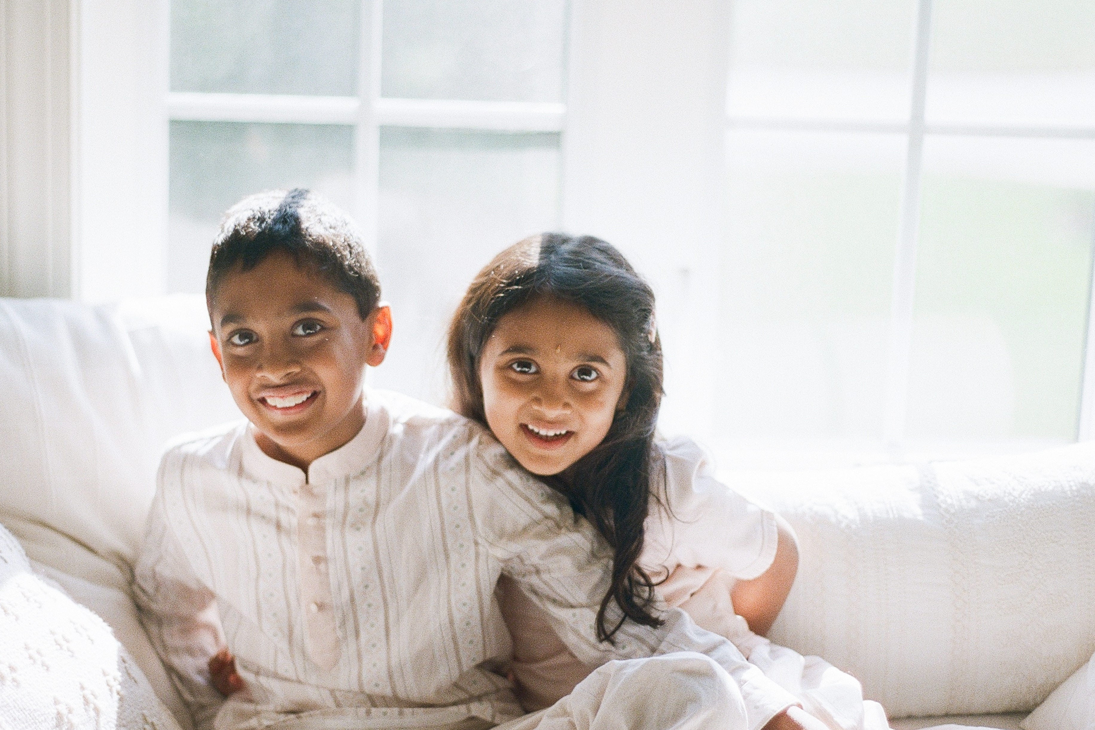 Two children sitting on a white couch in a bright room with large windows.