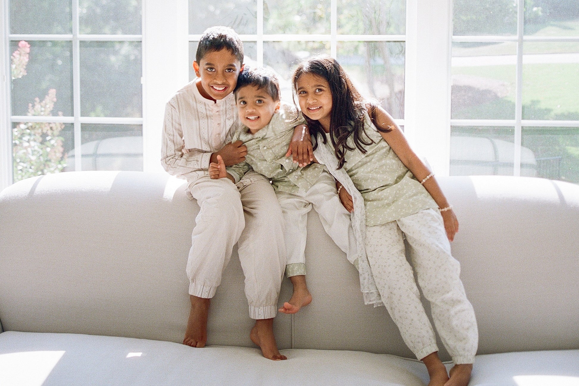 Three children sitting on a white couch in a bright room with large windows.